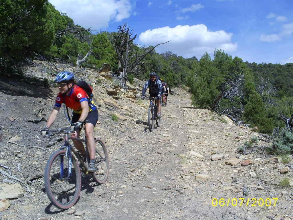 A group of mountain bikers riding on a rocky trail surrounded by lush greenery and scattered trees under a partly cloudy sky. The foreground features a cyclist in a red and blue jersey, while two others can be seen in the background on the trail. Paradox Trail mountain bike trail.