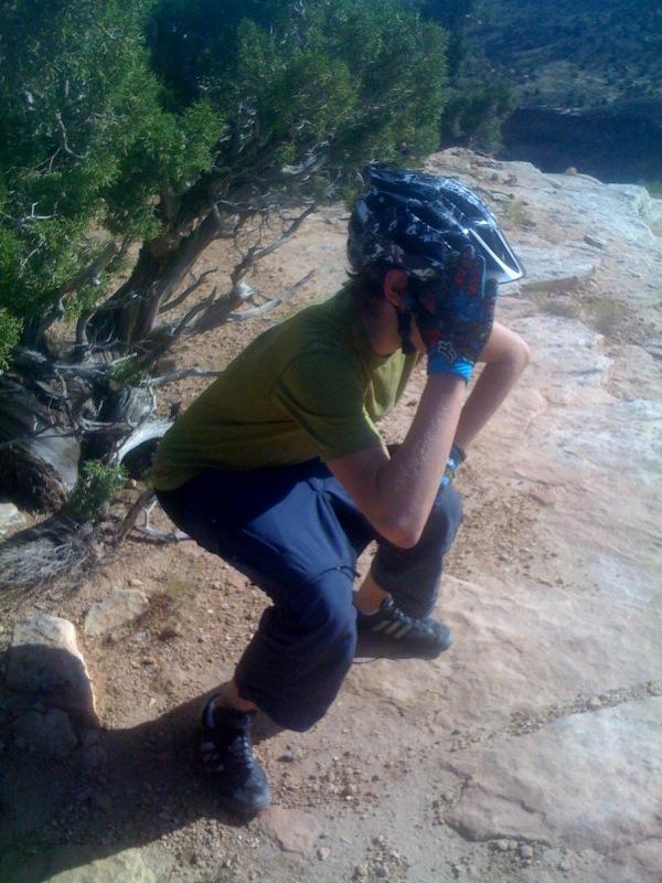 A person squatting on rocky terrain, wearing a cycling helmet and gloves, with one hand covering their face. Surrounding them are sparse shrubs and a scenic background of hills or cliffs. The scene suggests outdoor activities such as mountain biking or hiking. Mary's Loop / Horsethief Bench mountain bike trail.