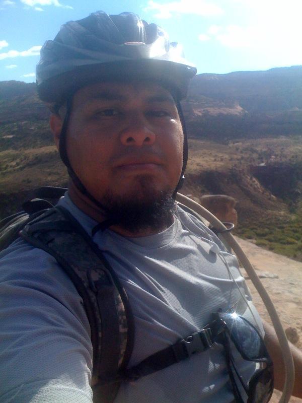 A man wearing a bicycle helmet and a gray athletic shirt, with a backpack, poses for a selfie against a scenic outdoor background featuring hills and blue sky. Mary's Loop / Horsethief Bench mountain bike trail.