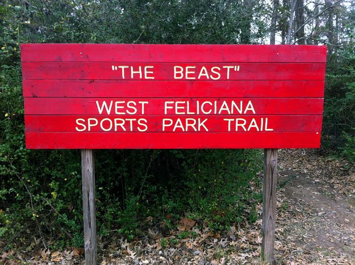 A bright red wooden sign reading "THE BEAST" and "WEST FELICIANA SPORTS PARK TRAIL," surrounded by trees and foliage. The Beast mountain bike trail.