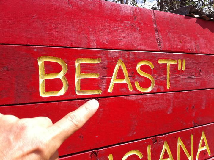 A close-up image of a red wooden sign with the word "BEAST" carved into it, highlighted in yellow. A person's hand, pointing towards the word, is visible in the foreground. The background features trees and a blue sky. The Beast mountain bike trail.