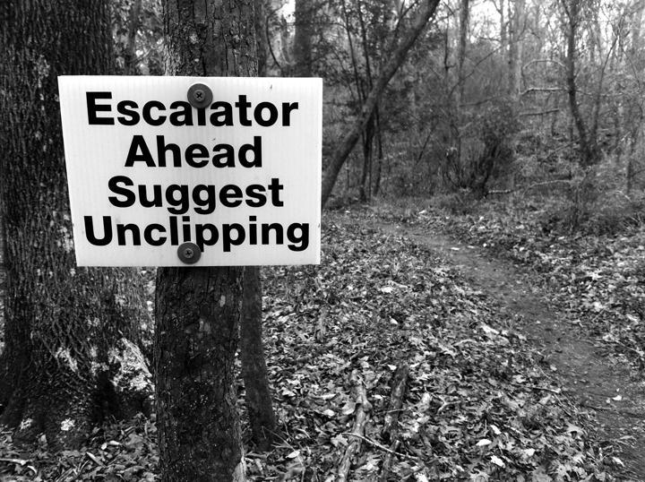 A black and white photograph of a sign attached to a tree in a forested area. The sign reads, "Escalator Ahead Suggest Unclipping," in bold text. The background features a dirt path winding through the trees and scattered fallen leaves on the ground. The Beast mountain bike trail.