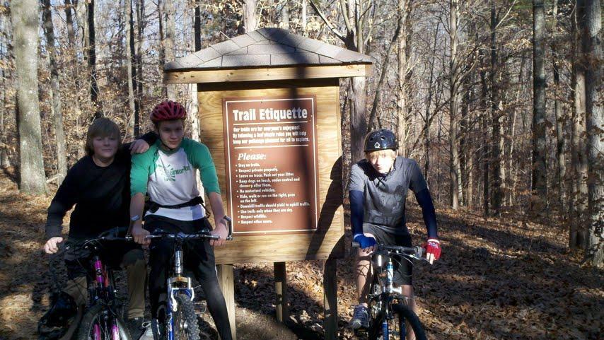 Three children are standing next to their mountain bikes in front of a wooden sign that displays trail etiquette guidelines. The scene is set in a wooded area during daytime, with autumn leaves scattered on the ground. The sign provides important tips for trail users, ensuring safety and respect among bikers and hikers. Rotary Park mountain bike trail.