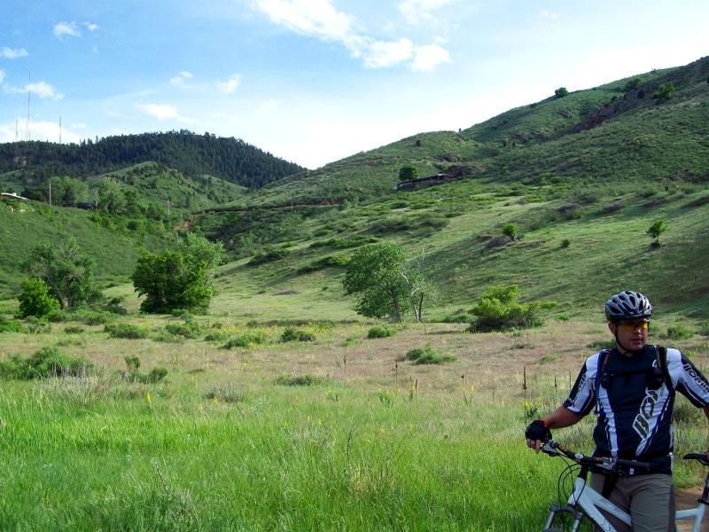 A mountain biker standing beside his bike in a lush, green landscape with rolling hills and trees under a clear blue sky. The scene is vibrant, showcasing the natural beauty of the outdoors. Chimney Gulch mountain bike trail.