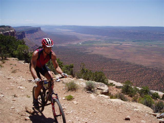 A mountain biker riding on a rocky trail, surrounded by scenic views of a vast, arid landscape with distant hills and a river. The sun is shining brightly in a clear blue sky. Paradox Trail mountain bike trail.