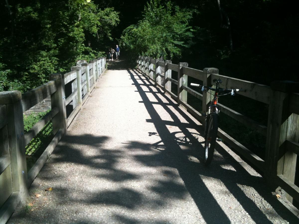 A sunlit pathway through a wooded area, featuring a wooden railing alongside a gravel path. A parked bicycle is visible on the right side of the path, with shadows of the railing casting patterns on the ground. In the distance, two people can be seen walking along the path. Wissahickon Valley Park mountain bike trail.