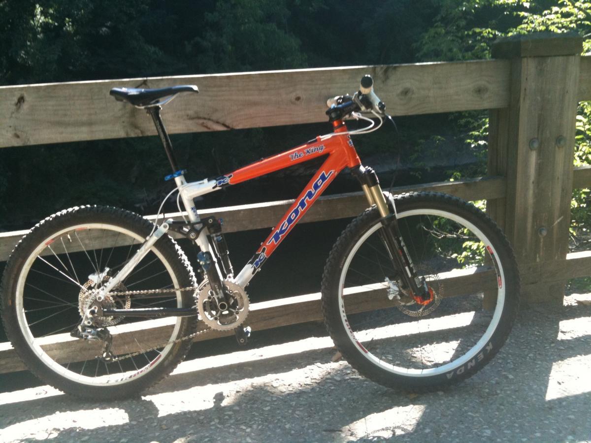 A mountain bike with a vibrant orange and white frame is resting against a wooden railing. The bike features thick tires suitable for off-road trails and has a visible gear system and suspension fork. The background includes greenery and a glimpse of water, suggesting a scenic outdoor location. Wissahickon Valley Park mountain bike trail.