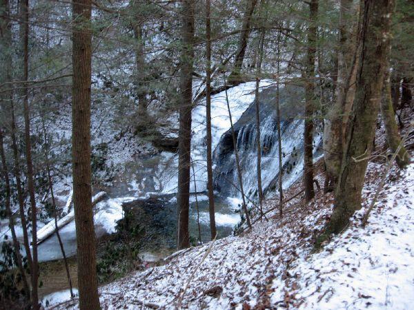 A serene winter landscape featuring a small waterfall surrounded by snow-covered rocks and trees. The water flows gently over the rocks, while the ground is blanketed with snow, creating a tranquil, nature-filled scene. The Bee Trail mountain bike trail.