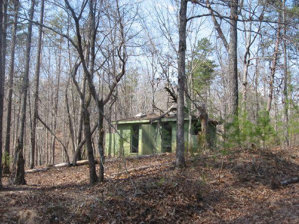 A small, abandoned green structure nestled among bare trees in a wooded area, with fallen leaves on the ground and a clear blue sky above. The building appears weathered and is partially obscured by the surrounding foliage. Blackburn State Park mountain bike trail.