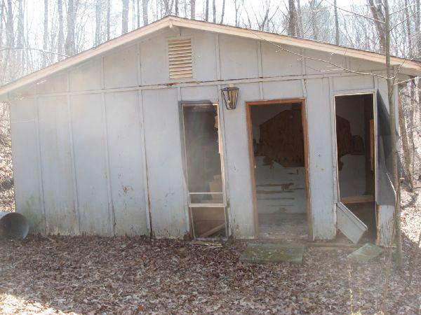 An abandoned, weathered building situated in a wooded area, with several doors ajar and peeling paint on the exterior. The surrounding ground is covered with fallen leaves, and trees loom in the background. Blackburn State Park mountain bike trail.
