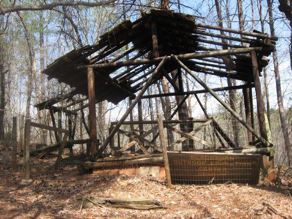 A dilapidated wooden structure surrounded by trees in a forested area. The roof is partially collapsed, and the walls are made of logs with some fencing visible. Leaves and debris cover the ground around the structure, indicating abandonment. A sign on the structure reads "OUTDOOR ENCLOSURE." Blackburn State Park mountain bike trail.