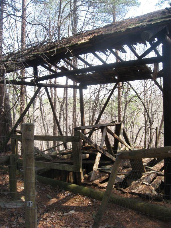 An abandoned wooden structure partially covered by foliage, with a weathered roof and exposed beams. In the foreground, there is a broken fence leading to a dark opening below. Surrounding the structure is a forest of bare trees, indicating it is situated in a remote, wooded area. Blackburn State Park mountain bike trail.