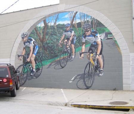 A colorful mural depicting three cyclists riding on a scenic road surrounded by trees and greenery. The mural is painted on a concrete wall and features a vibrant blue sky in the background.
