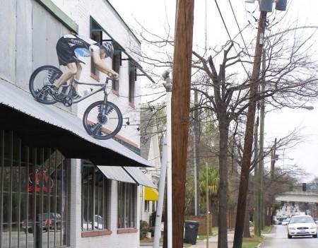 A mural of a cyclist riding a mountain bike is painted on the side of a building, appearing to leap off the wall. The scene is set on a cloudy day with trees and utility poles in the background, and a street with a parked car is visible.