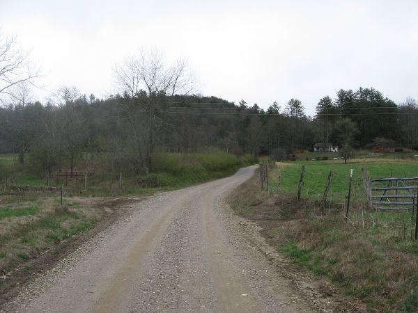 A winding gravel road leads through a rural landscape, flanked by bare trees and grassy fields. In the background, a small house is nestled among the trees on a gentle hill, under a cloudy sky. Fencing lines the road, adding to the tranquil countryside scene. Canada Creek mountain bike trail.