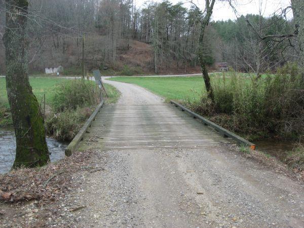 A wooden bridge spans a small creek, leading onto a gravel road that curves gently into the distance. Lush green grass surrounds the area, while trees line the background, indicating a rural setting. The sky is overcast, creating a calm and serene atmosphere. Canada Creek mountain bike trail.