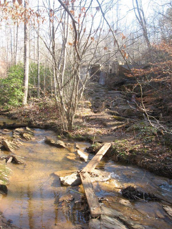 A peaceful scene of a woodland creek surrounded by trees, with a rocky bank and a wooden plank crossing over the water. In the background, a small waterfall is partially visible, suggesting a gentle flow of water. The foliage is sparse, indicating it may be early spring or late autumn. White Tail mountain bike trail.