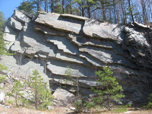 A large, weathered rock face featuring horizontal layers of stone, surrounded by sparse vegetation and tall trees in the background. The rock exhibits distinct, flat edges and varying shades of gray, with patches of underbrush and small pine trees at its base. Rock Quarry Road mountain bike trail.