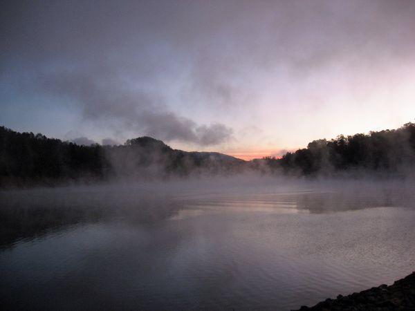 A serene early morning scene featuring a misty lake surrounded by hills. The water reflects the muted colors of dawn, with soft pink and blue hues in the sky. Light fog hovers above the surface of the water, creating a tranquil and ethereal atmosphere. Tsali Recreation Area mountain bike trail.