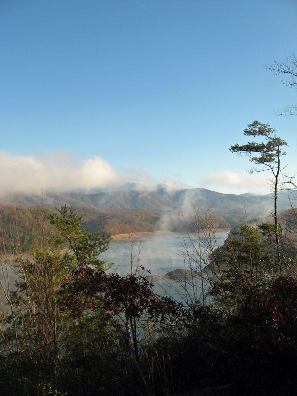 A scenic view of a lake surrounded by mountains, with mist rising from the water and trees in the foreground. The sky is clear with a hint of clouds in the background, creating a peaceful and serene atmosphere. Tsali Recreation Area mountain bike trail.