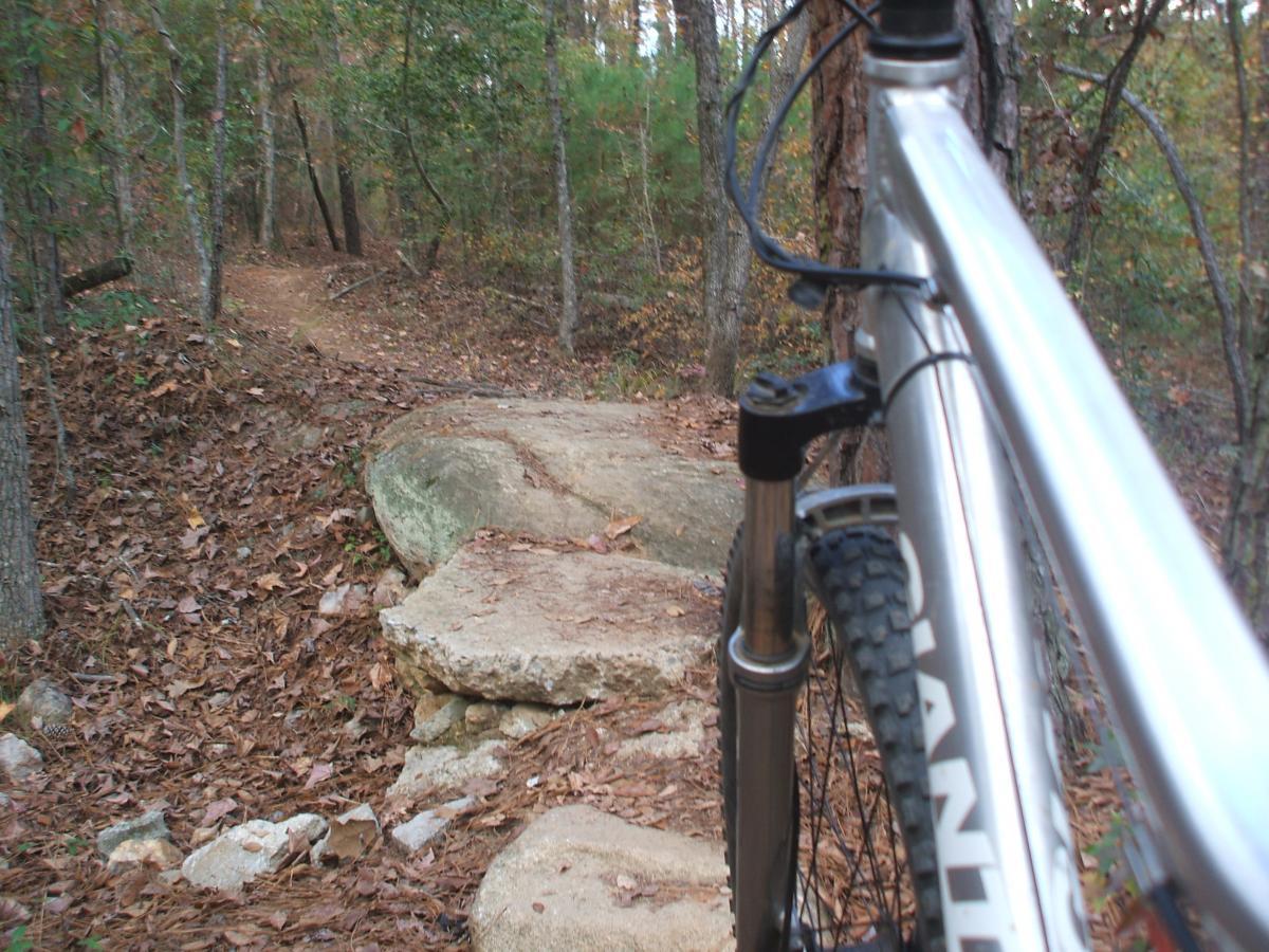 A mountain bike positioned at the edge of a rocky trail through a wooded area, with fallen leaves covering the ground and a winding path visible in the background. Great Wall Loop mountain bike trail.