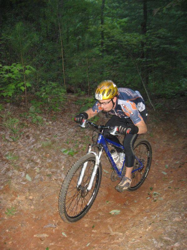 A mountain biker navigates a dirt trail in a forested area, wearing a yellow helmet and cycling gear. The cyclist leans forward on the bike, showing focus and intensity as they maneuver through the rugged terrain. Surrounding greenery includes trees and underbrush. Bull / Jake Mountain mountain bike trail.