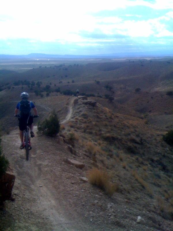 A mountain biker rides along a narrow dirt trail winding through hilly terrain, with scenic vistas of rolling hills and sparse vegetation in the background. The sky is partly cloudy, suggesting an outdoor adventure in a rugged landscape. Joe's Ridge mountain bike trail.
