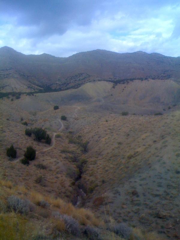 A panoramic view of a dry, mountainous landscape with rolling hills, sparse vegetation, and a winding dirt path. The sky is partly cloudy, casting a soft light over the terrain. Joe's Ridge mountain bike trail.
