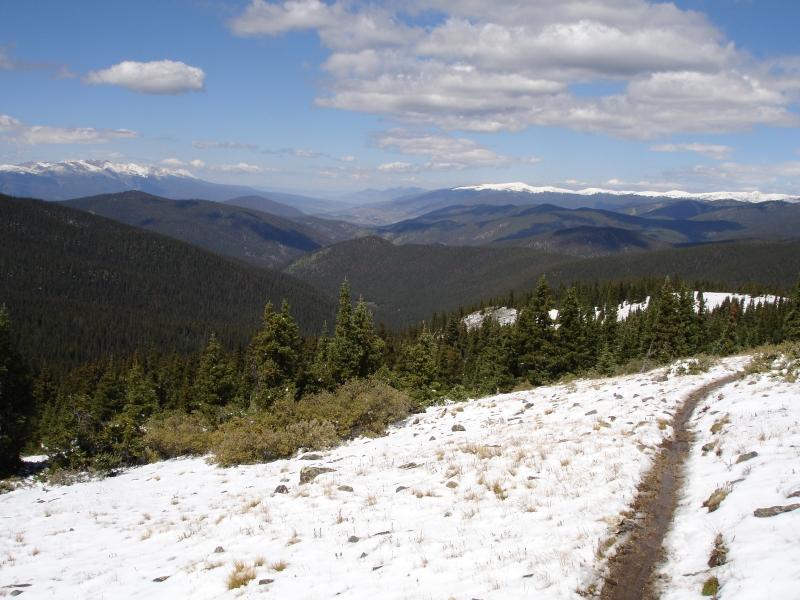 A scenic mountain landscape featuring snow-capped peaks in the distance, rolling green hills, and a winding dirt path. The foreground has patches of snow and rocky terrain, while expansive valleys and blue skies with fluffy white clouds fill the background. West Jefferson Trail mountain bike trail.