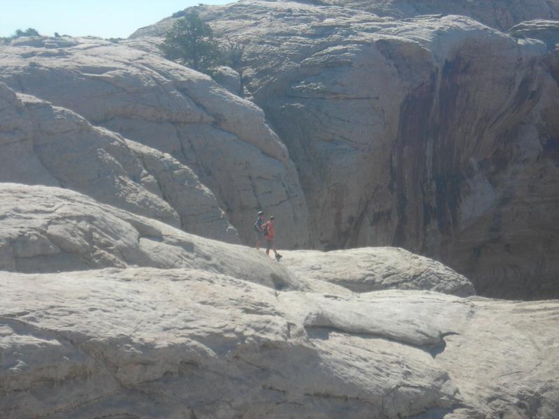 A hiker stands on a rocky outcrop surrounded by large, textured rock formations under a clear blue sky. The hiker is wearing a red shirt and appears to be exploring the rugged terrain. Klondike Bluffs mountain bike trail.