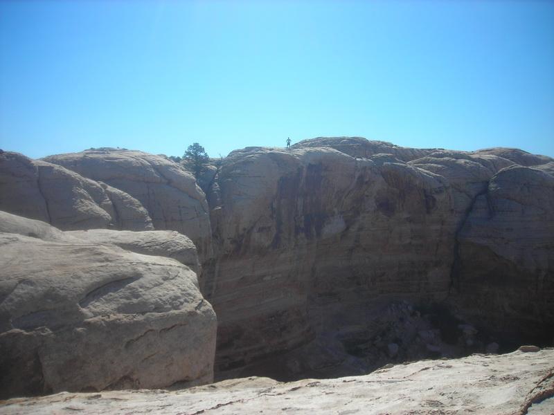 A rocky landscape under a clear blue sky, featuring rugged cliffs and a small figure standing on the edge of the rock formation. Klondike Bluffs mountain bike trail.