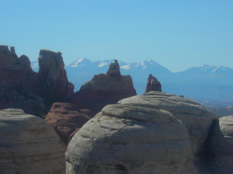A panoramic view of unique rock formations in a desert landscape, with large, rounded rocks in the foreground and towering, jagged cliffs in the background. Snow-capped mountains stretch across the horizon under a clear blue sky. Klondike Bluffs mountain bike trail.