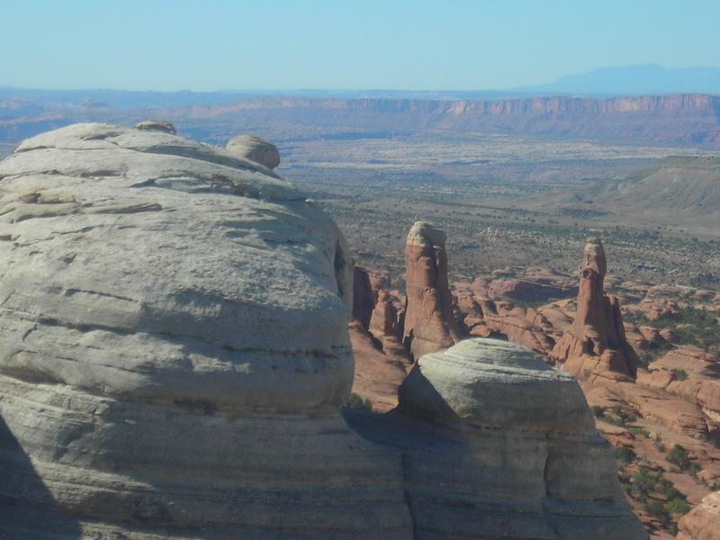 A panoramic view of a rocky desert landscape featuring unique rock formations. In the foreground, there are smooth, rounded boulders, while in the background, tall spire-like rock formations rise prominently against a clear blue sky, with layered cliffs and valleys stretching into the distance. Klondike Bluffs mountain bike trail.