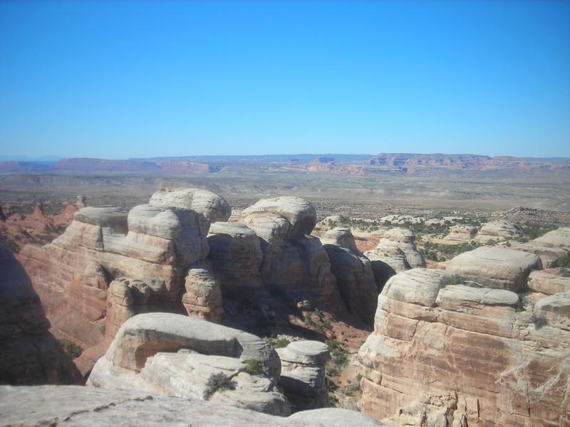 A panoramic view of rugged rock formations and distant mesas under a clear blue sky. The landscape features layered sandstone structures and sparse vegetation, showcasing the natural beauty of a desert environment. Klondike Bluffs mountain bike trail.