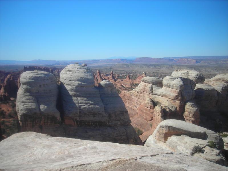 A panoramic view of towering rock formations in a desert landscape under a clear blue sky, with reddish-brown cliffs and distant mountains in the background. Klondike Bluffs mountain bike trail.