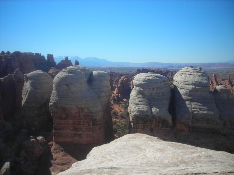 Rock formations stand prominently against a clear blue sky, showcasing a layered landscape of red and gray stone. In the background, distant mountains are visible on the horizon, adding depth to the scene. The focus is on the unique shapes of the rocks, with some appearing rounded and smooth while others rise sharply, highlighting the natural beauty of the area. Klondike Bluffs mountain bike trail.