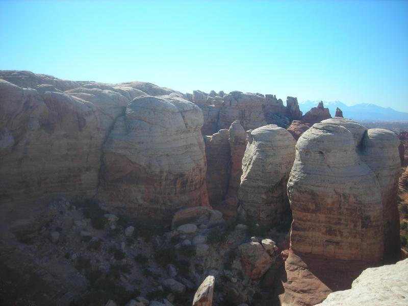 A scenic view of rocky formations in a desert landscape, showcasing layered sandstone cliffs and large rock spires under a clear blue sky. The foreground features rounded rock structures, while the distant background reveals rugged hills and mountains. Klondike Bluffs mountain bike trail.