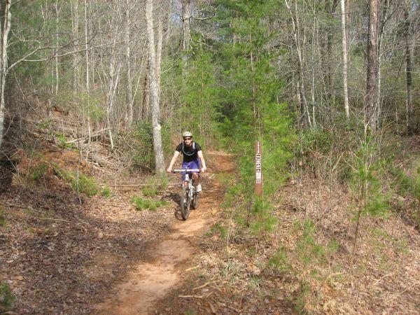 A mountain biker riding along a dirt trail surrounded by trees and greenery. A trail sign is visible alongside the path. No Tell Trail mountain bike trail.