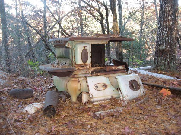 An old, rusty stove lies abandoned in a forested area, surrounded by fallen leaves and pine needles. The stove, with a faded green and white exterior, shows signs of wear and neglect, blending into the natural environment of trees and underbrush. Flint Ridge mountain bike trail.