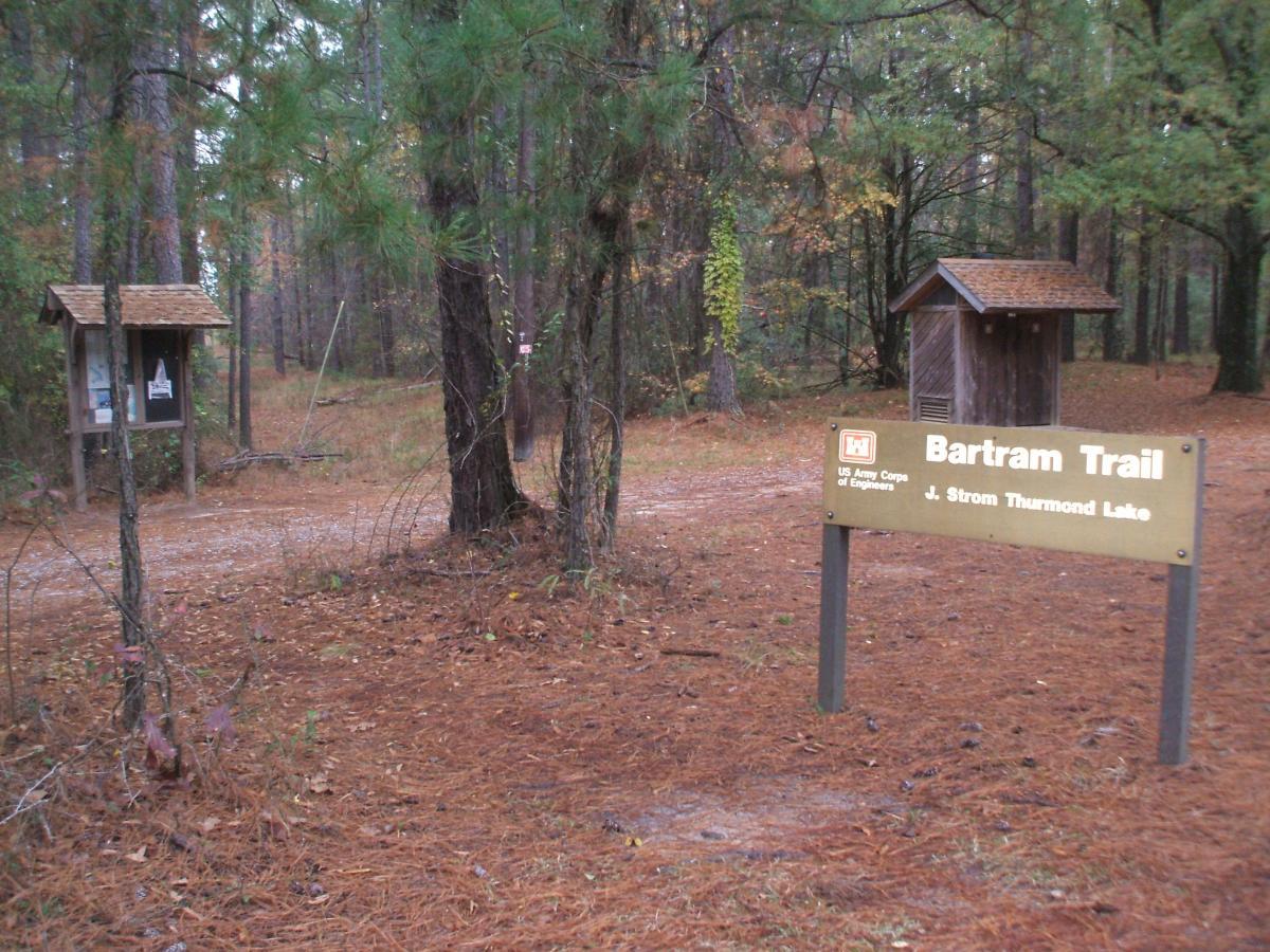 Sign for the Bartram Trail at J. Strom Thurmond Lake, situated in a forested area with pine trees and a dirt path. A trailhead information kiosk is visible on the left, and a small wooden structure is on the right. The ground is covered in pine needles, indicating a natural, serene environment. Bartram Trail / West Dam / Wildwood Park mountain bike trail.