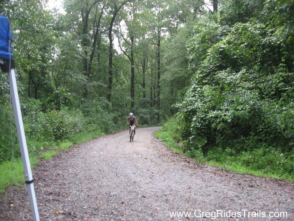 A cyclist riding on a gravel path through a lush, green forest on a cloudy day, surrounded by tall trees and dense foliage. Winding Stairs Loop mountain bike trail.