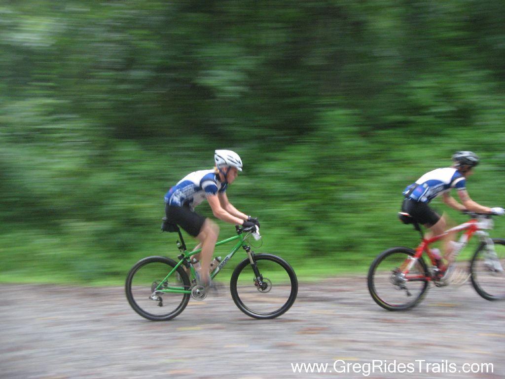 Two mountain bikers riding on a gravel trail with a blurred background, suggesting speed. One rider is on a green bike, while the other is on a red bike. The scene is set in a lush green environment, indicating a forested area. Winding Stairs Loop mountain bike trail.