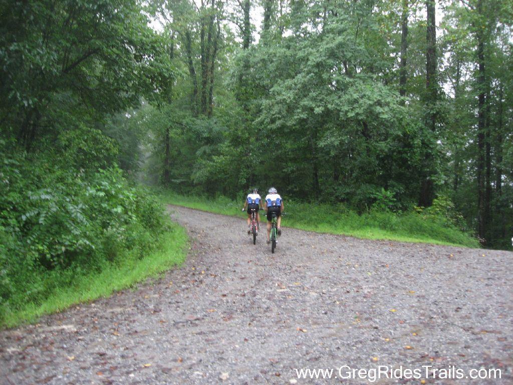Two cyclists riding on a gravel path surrounded by lush green trees in a misty, rainy environment. Winding Stairs Loop mountain bike trail.
