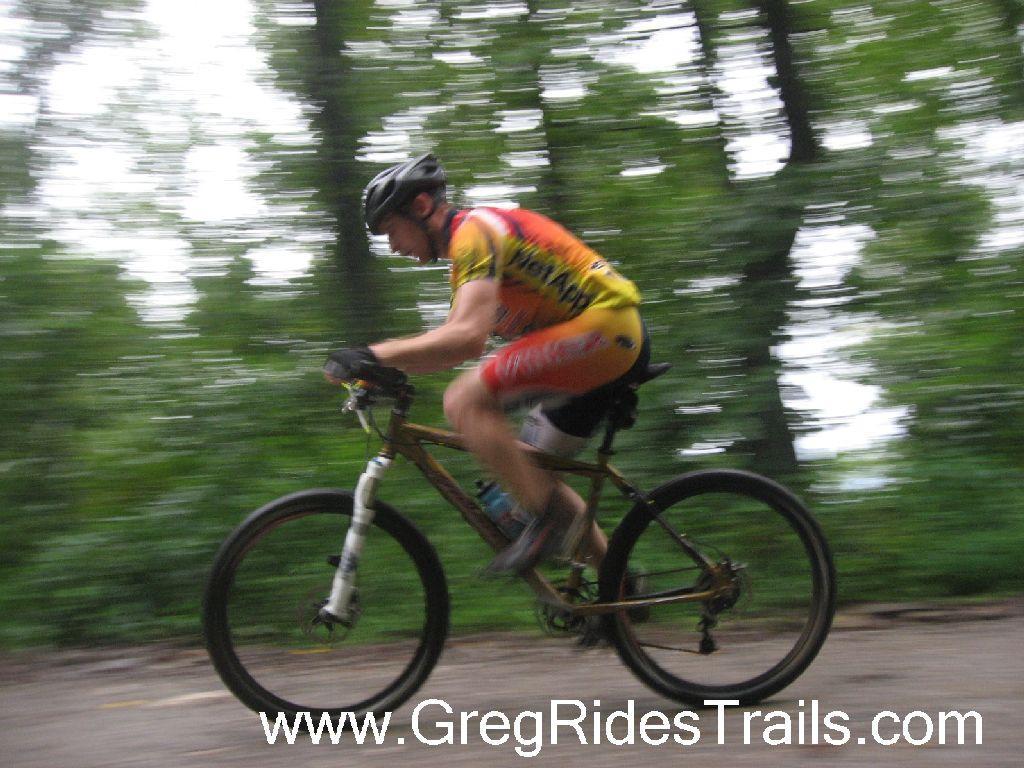 A cyclist in motion riding a mountain bike on a wooded trail, captured with a blur effect to emphasize speed. The rider is wearing a colorful jersey and helmet, surrounded by lush green trees in the background. Winding Stairs Loop mountain bike trail.