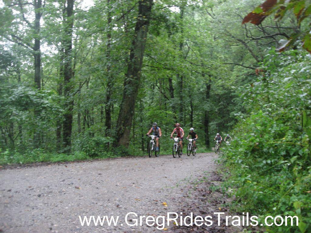 A group of mountain bikers riding on a gravel trail through a lush, green forest after rain. Tall trees line the path, creating a natural canopy, while the terrain appears muddy and wet, indicating recent rainfall. The cyclists are in various positions, showcasing their biking gear and enjoying the ride. Winding Stairs Loop mountain bike trail.