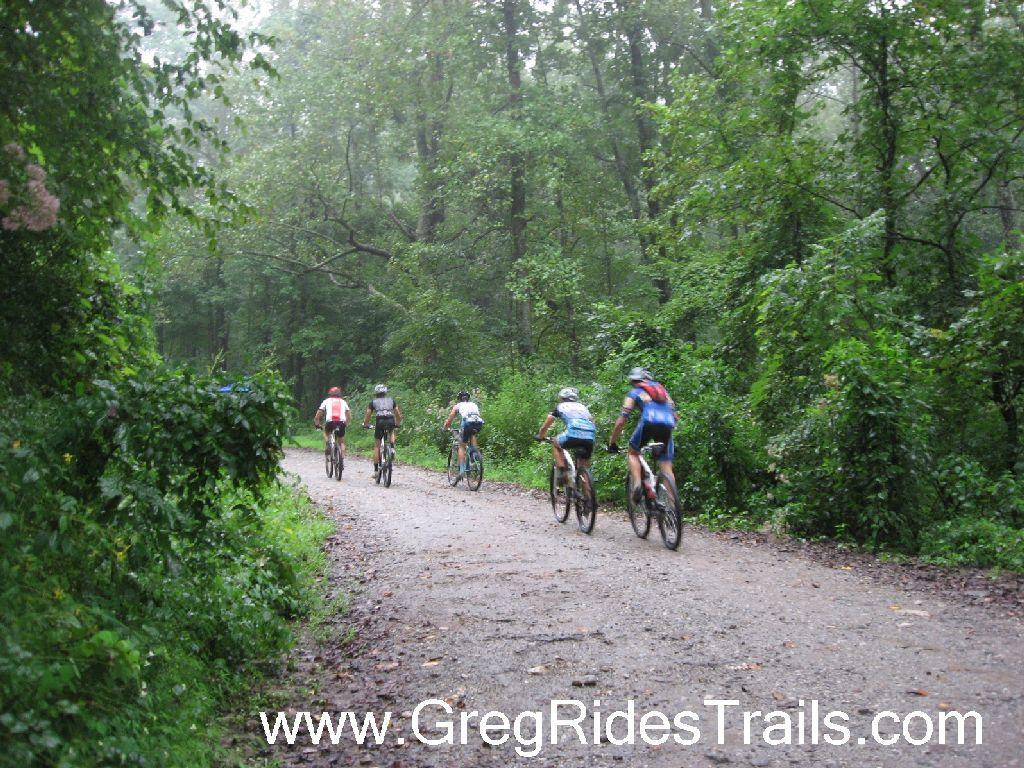 A group of five mountain bikers riding along a muddy trail surrounded by lush green foliage in a forested area, under overcast skies. Winding Stairs Loop mountain bike trail.
