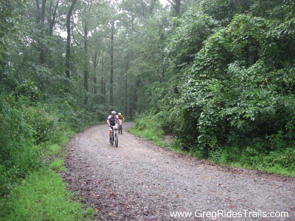 Two mountain bikers ride along a gravel trail surrounded by lush green forest under rainy weather. The scene captures the essence of outdoor adventure and camaraderie in a natural setting. Winding Stairs Loop mountain bike trail.