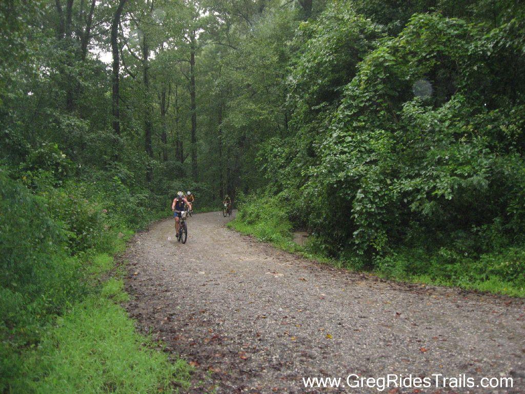 A group of cyclists navigating a gravel trail through a lush, green forest on a rainy day. The path is surrounded by tall trees and dense foliage, creating a vibrant and natural atmosphere. Winding Stairs Loop mountain bike trail.
