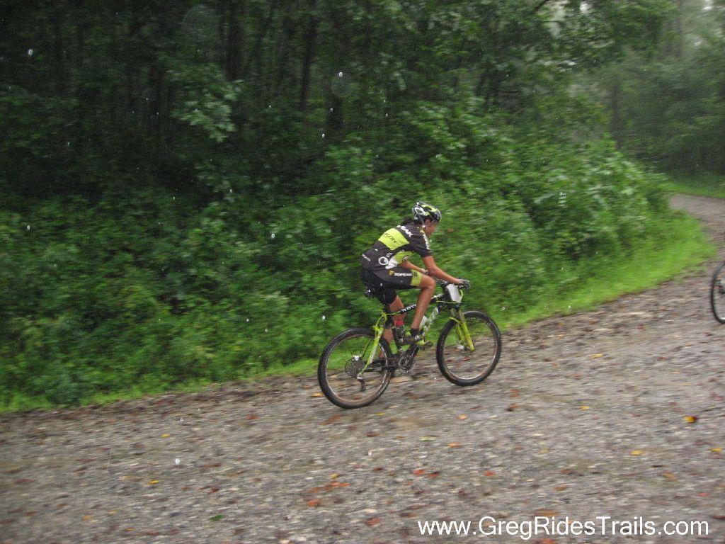 A cyclist riding a mountain bike on a gravel path through a wooded area, with rain falling and greenery surrounding the trail. Winding Stairs Loop mountain bike trail.