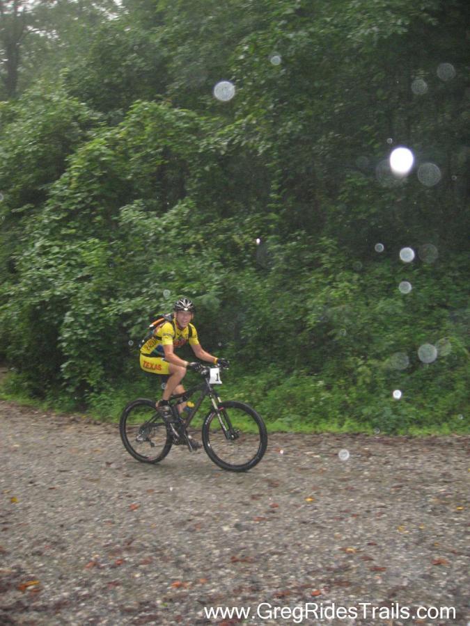 A cyclist wearing a yellow jersey is riding a mountain bike on a gravel path, surrounded by dense greenery. The scene appears to be rainy, with water droplets visible in the air and on the ground. The cyclist is focused and in motion, showcasing determination despite the wet conditions. Winding Stairs Loop mountain bike trail.